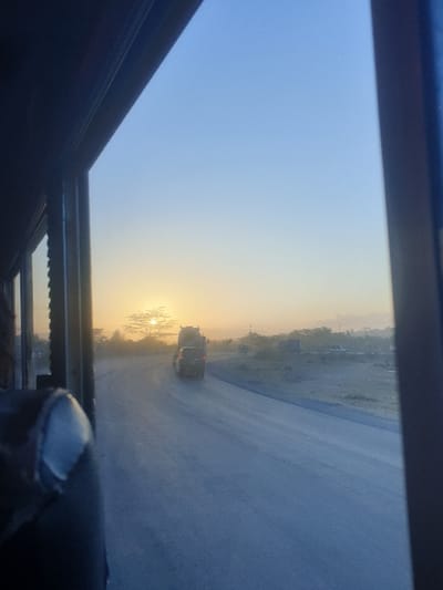 The image captures a view from inside a vehicle, looking out onto a road scene during either sunrise or sunset. The perspective is from a window or possibly a sunroof, indicated by the dark framing visible on the top, bottom, and partially on the left and right edges. The image is rotated counter-clockwise, making the horizon appear slanted.

The sky dominates the upper portion of the frame, transitioning from a soft, light blue in the top left to a brilliant golden-yellow and orange glow on the right, where the sun is low on the horizon. This strong backlighting silhouettes elements closer to the sun.

A paved road or highway stretches into the distance, occupying the mid-ground of the image. It appears somewhat dusty or hazy, especially as it recedes towards the horizon. On the left side of the road, a barren or sparsely vegetated landscape with indistinct distant structures or hills is visible. On the right side, closer to the bright sun, a cluster of trees or bushes stands out as dark silhouettes against the intense light.

Two vehicles, likely cars or SUVs, are visible on the road in the mid-distance, heading away from the viewer. They also appear as dark silhouettes against the bright backdrop, making their specific details indistinguishable. The time of day is clearly marked by the low sun and the warm, golden hues illuminating the scene, characteristic of either early morning (sunrise) or late afternoon/evening (sunset). The overall atmosphere has a hazy quality. Part of the vehicle's interior, possibly a hand or gloved finger resting on the window frame, is visible in the bottom right corner of the frame.