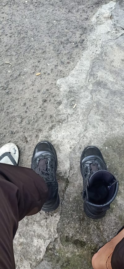 This media file depicts a close-up, high-angle shot of a person's feet and lower legs, positioned on a rough, grey concrete surface. The setting appears to be outdoors, given the texture of the ground and the presence of what look like discarded cigarette butts scattered on the concrete. The concrete itself is uneven and shows signs of wear and cracking.
The primary subjects are the person's footwear. On the left, a person is wearing dark-colored pants and a white flip-flop. To the right, the person is wearing two black tactical boots. One boot is fully on, showing a lace-up system with a quick-lace closure mechanism and a side zipper. The other boot is partially unzipped and appears to be slightly loosened, revealing the inner lining. The contrast between the casual flip-flop and the sturdy tactical boots suggests a mix of activities or perhaps a transition between different environments.
There are no people fully visible in the frame, only parts of their legs and feet. The lighting suggests it might be daytime, possibly overcast, as there are no strong shadows. The overall mood is neutral, focusing on the practical elements of footwear and the environment. The location cues of "Tambontunong, Indonesia" are provided externally and are not visually discernible from the image itself, beyond the general impression of an outdoor, possibly urban or semi-urban, environment. There is no visible text within the image.