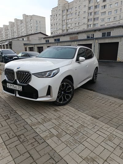A white BMW X1 is parked on a wet brick-paved surface in front of a row of garages. The garages have dark brown doors and are attached to a low-rise brick building. Behind the garages are tall, multi-story apartment buildings, characteristic of urban residential areas. The sky is overcast, suggesting a cloudy or rainy day, with raindrops visible on the car's surface. The license plate of the BMW reads "O 111 KC 31 RUS", indicating it is registered in Russia. The overall scene depicts a residential area in the city of Gubkin, Russia, with a focus on the modern vehicle and its surroundings.