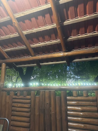 The image depicts a rustic wooden structure with a red tiled roof, likely a gazebo or covered outdoor seating area, situated in the city of Arinj, Armenia. The structure features rough-hewn wooden beams supporting the roof and a fence made of horizontally stacked logs. String lights are visible, creating a festive ambiance against a backdrop of lush green trees and a twilight sky. The overall impression is one of a natural, relaxed setting, possibly a restaurant or a recreational space.