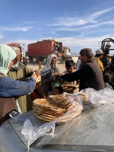 An outdoor scene in Chaman City Tehsil, Pakistan, depicts a large gathering of individuals, predominantly adults and children, on dry, unpaved ground under a clear blue sky with sparse clouds. In the foreground, stacked round flatbread (likely naan) is visible on the hood or roof of a light-colored vehicle, partially covered by clear plastic bags.
Several individuals are engaged in food distribution. On the left, a person wearing a light blue head covering and garment receives a piece of flatbread from an older man with a white beard. To the right, a man with a dark beard and patterned cap, wearing a black vest, dispenses flatbread and small plates of yellow-colored food (possibly rice) to multiple recipients, including a young boy. Another man with a white head covering and beard holds a piece of flatbread.
In the mid-ground, numerous people, including children, observe the food distribution. A child in an orange garment is visible running in the background. Two large trucks, each carrying a red shipping container, are prominently parked in the background. These containers are heavily overloaded with bundled personal belongings and household items, suggesting a context of movement or displacement. One truck has a visible framework or ladder attached to its rear. A light-colored tent structure is also visible in the mid-background. Distant mountains define the horizon.