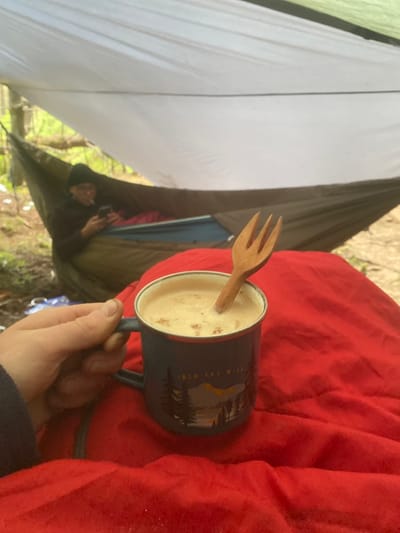A person is holding a blue mug filled with a light brown liquid, possibly coffee or tea, and a wooden fork, with a red blanket underneath. The mug has a design on it that says "Into the Wild" with a mountain scene. The person is in a hammock, under a white tarp with a person sitting in the hammock behind them, out of focus. They are in Macclesfield Forest in the United Kingdom, at coordinates 53.26 latitude and -1.991 longitude. The scene is in the woods, possibly in a campsite.