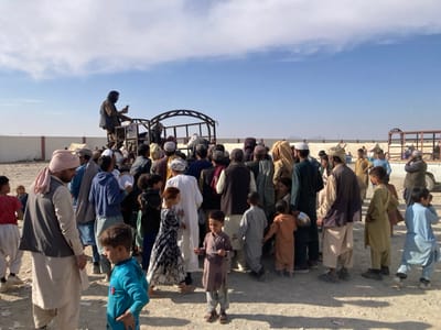 An outdoor scene in Chaman City Tehsil, Pakistan, depicts approximately 50-70 individuals, primarily adult men and children of various ages, gathered on dry, unpaved ground under a clear blue sky. A white pickup truck with a metal frame canopy serves as a focal point, with people clustered densely around its rear. One man is elevated on the truck's bed, holding a small rectangular object, potentially a mobile phone, and appears to be addressing or observing the crowd. Numerous individuals in the gathering, including many children, are holding white plates or bowls. In the background, a light-colored wall runs horizontally, behind which white tents and distant, hazy terrain features are visible. The environment is arid, with strong shadows indicating direct sunlight.