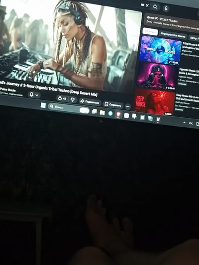 An overhead medium shot captures a laptop screen displaying a YouTube video featuring a female DJ with braided hair wearing headphones and tribal attire, operating a DJ console. To the right of the main video, a sidebar showcases recommended videos with titles like "Tribal House & Psychedelic", "Hypnotic House | Deep Music & Atmosphere", and "Deep House Mix | Late Night Chill and Smooth Beats." Below the main video, a search bar with the inscription "Поиск" (Search) is visible, along with YouTube interface elements such as like buttons, share icons, and a subscribe notification bell. The laptop is placed on a dark surface, with a portion of a person's feet in the foreground, suggesting the viewer is sitting or lying down while watching. The background is predominantly dark and out of focus, with hints of ambient light. The provided image does not contain any elements that would allow for the determination of the location within Krasnoarmeysk, Russia.