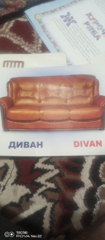 This image showcases a brown leather sofa with three cushions. The sofa has a tufted design and appears to be in good condition. Below the sofa, text is visible in two languages, likely indicating "sofa" in Russian and English. The word "ДИВАН" is written in blue Cyrillic letters, while "DIVAN" is written in red Latin letters.
In the background, a children's book is partially visible. The book has a decorative border and a title written in a large, stylized font. Some of the text in the book appears to be in Russian, with words like "КУРОЧКА" and "РЯБА" being discernible, suggesting it might be a fairy tale. There's also a small circular image with the number "8" on the book's page.
The overall setting seems to be indoors, possibly a living room, given the presence of the sofa and the carpet in the foreground, which features a reddish-brown and beige pattern. The lighting suggests it could be daytime. A faint "TECNO POVA Neo 2" logo is visible in the bottom left corner, indicating the device used to capture the image.