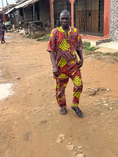 A man stands in a dirt street in Ijegun, Nigeria. He wears a patterned shirt and pants, and sandals. Behind him is a building with barred windows. To the left is a shop. The street is wet.