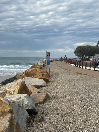 The image depicts a scene at Muttonbird Island Nature Reserve in Coffs Harbour, Australia, characterized by a cloudy sky over the ocean. In the foreground, a rocky breakwater composed of large, rough-hewn stones lines the edge of the water. To the right of the breakwater, a paved pathway runs parallel to the coast. Two individuals are walking away from the camera on this pathway. Beyond the pathway, a road is visible with several cars. Mature trees with dense foliage are situated further inland. A signpost with warning information is positioned on the left side of the pathway near the breakwater. The environment suggests a coastal, potentially breezy, day.