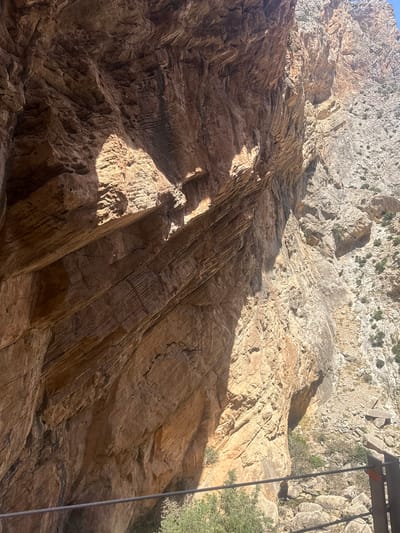 This media file captures a dramatic view of a rugged, rocky cliff face in Ardales, Spain. The scene is dominated by the textured geological formations, with layers of rock displaying varying shades of brown and grey. Sunlight dramatically illuminates parts of the cliff, creating strong contrasts with deep shadows. The rock surfaces are rough and uneven, showing signs of erosion and fissuring, with some areas appearing more weathered and porous than others.

The perspective is looking upwards towards the top of the cliff, suggesting the viewer is at the bottom or in a canyon. In the upper right of the image, a portion of a cable or wire is visible, suggesting the presence of a man-made structure, possibly related to a cable car, zipline, or a walkway suspended along the cliff face. A hint of green foliage is also visible, clinging to the upper parts of the cliff and in the background, indicating some vegetation in this arid environment.

The time of day appears to be mid-morning or late afternoon, judging by the angle and intensity of the sunlight casting long shadows across the rock. The weather seems clear and sunny, contributing to the stark lighting conditions. There are no people or animals directly visible in the frame. The overall impression is one of natural grandeur and a sense of scale, emphasizing the raw power of geological processes. The textures and lighting evoke a feeling of awe and perhaps a touch of adventure, hinting at outdoor exploration activities. No text is visible in the image.