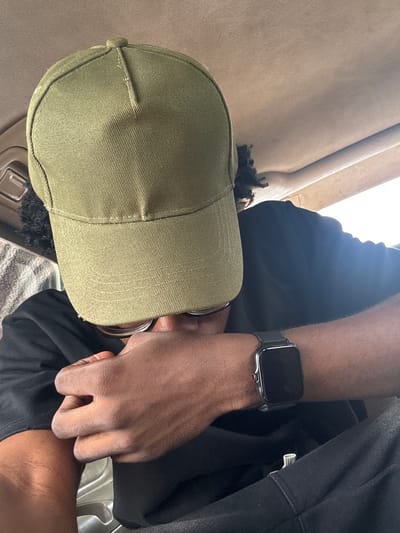 The image shows a person from the chest up, wearing a green baseball cap and a black t-shirt. They are also wearing a smartwatch on their left wrist. The person's hands are visible, and one hand appears to be resting on their lap. The background suggests an indoor setting, possibly a car or a room with light-colored upholstery. The lighting indicates it is daytime, with sunlight streaming in from the side. The scene is a close-up shot, focusing on the person's attire and accessories. There is no visible text in the image. The location context provided is Orji, Nigeria.