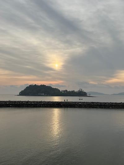The image captures a tranquil scene near Pak Nam Chumphon, Thailand, showing a sunset reflecting on the water. A long, dark rock barrier extends into the water, with the reflection of the setting sun creating a bright, shimmering effect on the surface. In the distance, a lush green island rises from the sea, partially obscuring the horizon. The sky is filled with soft, muted colors, blending pale gray clouds with hints of orange and yellow from the setting sun. Two figures are visible on the rock barrier, appearing small against the vastness of the landscape. Further out, a small boat can be seen near the island.