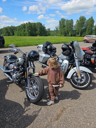 The image shows a child in a brown tracksuit standing next to a black and chrome motorcycle on an asphalt surface. The motorcycle is equipped with a skull-themed fuel tank design and a black helmet rests on the seat. Two other motorcycles are parked nearby, including a silver one. In the background, there's a green field bordering a tree line beneath a blue sky with scattered clouds. The location is presumably near Borodinskogo Muzeya, Russia.