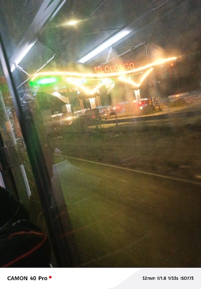 A nighttime view through a window captures the "MLOLONGO" illuminated sign above a tollbooth. Headlights and taillights of vehicles are visible in the background. The foreground shows a road surface and the blurred interior of what appears to be a bus. The photo is grainy, likely due to low light conditions and high ISO, taken with a "CAMON 40 Pro" at 32mm, f/1.8, 1/33s, and ISO773.