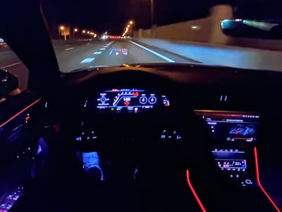 The image captures a nighttime view from the driver's seat of a car on a highway, possibly near Bezden, Bulgaria. The dashboard displays illuminated digital readouts, including a speed of 148, and a head-up display (HUD) projects speed and navigation information onto the windshield. Red and blue ambient lighting accents the interior. The road ahead is illuminated by streetlights and the car's headlights, revealing lane markings and the edge of the road. The car appears to be in motion.