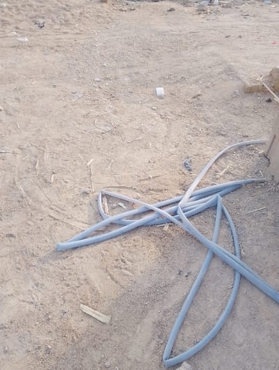 The image displays an outdoor ground scene, dominated by sandy and dusty terrain. The primary subject is a light blue, flexible hose or pipe, coiled and spread across the lower right and central portions of the frame. The hose appears to have a slightly textured or woven surface.
The ground is a muted beige-brown, indicative of dry earth, and shows minor irregularities and scattered small debris. Among the debris are small pieces of wood, visible towards the bottom left and near the hose. A small, white square object, possibly plastic or paper, lies on the ground towards the upper middle of the frame. Faint impressions or tracks are visible on the sandy surface.
In the background on the far right, a partial view of a light-colored, possibly concrete or stone structure is visible, suggesting a construction or undeveloped area. The overall lighting indicates daytime, with bright, natural illumination casting soft or no discernible shadows. The weather appears clear and dry. No individuals are present in the scene, and no specific activity is occurring. There is no visible text within the image. The setting is consistent with an outdoor, potentially utilitarian or undeveloped area in Panshekara, Nigeria.