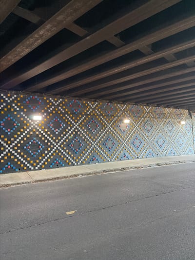 The photograph, taken in Atlanta, United States of America, presents a view looking down into a narrow passageway beneath what appears to be a bridge or overpass. On the left is a steel structure composed of intersecting beams and on the right is a gray asphalt surface, likely a road. Between the steel structure and the road is a narrow wall adorned with a geometric pattern of colored squares, primarily white, yellow, blue, and red. Three illuminated lights are positioned along this wall, their placement suggesting a pattern or design element. The overall composition is a study of urban infrastructure and the intersection of design, structure, and transportation.