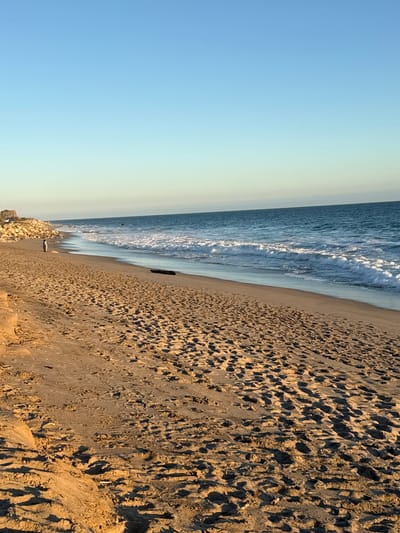 The image depicts a beach scene in Malibu, California, USA. The foreground is dominated by sand covered in numerous footprints, with some areas showing slight undulations and shadows. In the mid-ground, a solitary person stands near the water's edge, facing the ocean. A dark, elongated object, possibly a piece of driftwood, lies on the sand further down the beach. The ocean itself is calm with gentle waves breaking on the shore. The water transitions to a deep blue, meeting the horizon. In the background, a rocky shoreline extends to the left, with some structures visible on top. The sky is a clear, gradient blue, transitioning from a pale yellow near the horizon to a deeper blue overhead.