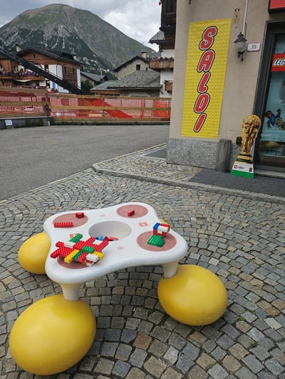 A bright yellow banner with red lettering, spelling out "SAVOIA", is displayed on a light-colored wall. The banner is adorned with a pattern of yellow circles. Above the banner, a small sign with the number "35" is visible, next to a decorative lamp. To the right of the banner, a tall, wooden sculpture shaped like a paintbrush, constructed from Lego bricks, stands prominently.
On a cobblestone pathway, a white Lego-themed activity table is set up. The table features red circular indentations and has Lego bricks in various colors, including red, green, and white, assembled on its surface. Two large, yellow, egg-shaped objects are attached to the base of the table, resembling wheels. The surrounding area is paved with gray cobblestones.
In the background, a picturesque mountainous landscape unfolds under a cloudy sky. Wooden buildings with dark roofs are nestled among the mountains. Orange construction netting is visible along a pathway. The scene appears to be outdoors in a village setting. The overall impression is one of a playful and welcoming environment, likely a Lego store or a public space featuring Lego-themed attractions.