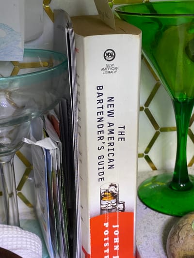 The image depicts a countertop scene in Ridgefield, United States. A white-spined book titled "THE NEW AMERICAN BARTENDER'S GUIDE" by John F. Poiste, published by New American Library, is prominently centered. Its spine features an illustration of a drink in a glass with ice. To the left, a clear glass margarita or cocktail glass with a light blue-green rim is partially visible, alongside other stacked papers or thin books and a segment of a white, textured cloth. To the right, a vibrant green stemmed glass, likely a martini or cocktail glass, stands on the countertop. The background consists of a wall covered in light-colored hexagonal tiles with subtle metallic or golden accents. The objects are situated on a light-colored countertop. No individuals or active interactions are present.