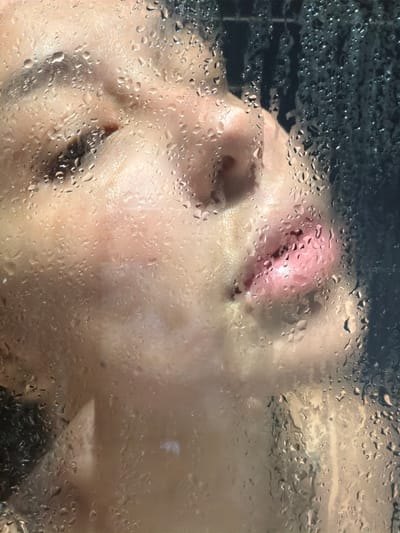 The image presents a close-up, intimate view of a person's face pressed against a transparent surface, heavily obscured by an array of water droplets and condensation. The droplets vary in size and density, causing significant distortion and blurring of the features behind.
The primary subject is a human face, characterized by a fair to light skin tone. Key features discernible through the condensation include the bridge and tip of a nose, and a partially open mouth revealing pink lips. On the upper right side of the face (viewer's left), the curve of a brow and a faint outline of an eye are visible, though the eye itself is largely indistinct. The person's expression appears subdued, possibly suggesting a moment of calm or introspection.
The transparent surface, likely glass from a shower door or steam room, acts as a visual filter. The myriad water droplets catch and reflect light, creating a sparkling, ethereal effect across the foreground. The background behind the person is dark and abstract, providing little detail but hinting at a confined, interior space.
The scene suggests the person is inside a shower or a steamy environment, such as a sauna or a bathroom, with their face close to the condensed barrier. The overall atmosphere conveys a sense of warmth, humidity, and a private, sensory experience. No discernible activity beyond the person's proximity to the glass is evident. There is no visible text within the image.