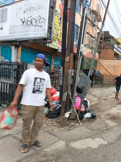 A man wearing a blue cap, white t-shirt, and khaki pants walks along a sidewalk carrying a red and green bag. He is positioned near a pile of trash bags. Behind him is a building with scaffolding and exposed brick. Metal poles stand along the sidewalk. Another person in a blue shirt and shorts can be seen further down the sidewalk.