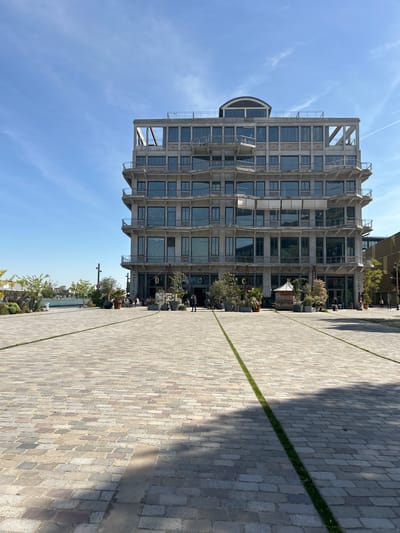 This media file captures an outdoor urban scene in Pantin, France, featuring a modern building next to a paved public space under a clear blue sky.

The prominent subject is a multi-story building on the left, characterized by its contemporary, industrial-chic design. It features a concrete facade with numerous large glass windows, exposed structural elements, and a distinctive curved window section near the top-left. The ground floor appears to host commercial establishments, with outdoor seating areas adorned with potted plants, small trees, and some decorative objects, including a small, light-colored, pointed-roof structure. Several people are visible here, some seated at tables, others standing, suggesting social activity or dining.

The foreground and right side of the image are dominated by a wide plaza paved with a mosaic of grey, beige, and reddish cobblestones. Two narrow, parallel strips of green grass run across this paved area, creating a neat geometric pattern. Long shadows are cast across the paving, indicating either morning or late afternoon sunlight.

The sky is bright and mostly clear, with a few faint wispy clouds and a subtle aircraft contrail.
Visible text on the building includes "MAGASINS" (French for "shops") in white letters, confirming the commercial nature of the ground floor. A green neon sign, possibly "BO" or similar, is also partially visible. The overall setting suggests a vibrant, possibly regenerated, urban area with a relaxed atmosphere.