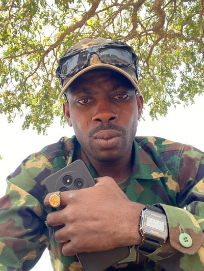 An adult male with a dark complexion is depicted in a close-up, low-angle shot, looking directly into the camera with a neutral expression. He wears a camouflage baseball cap with dark sunglasses resting on its brim, and a military-style camouflage jacket with visible green buttons on the cuff. A prominent gold-colored ring with an embossed design is on his left ring finger, and a black digital SKMEI watch with a dark metallic casing is on his right wrist. He holds a dark smartphone against his chest, displaying its rear camera module which features three distinct circular lenses/sensors. Visible above him are branches of a tree with numerous green leaves against a bright sky, indicating an outdoor setting. The scene is located in Zaki Biyem, Nigeria.