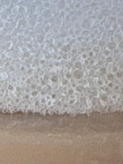 The image displays a close-up, top-down view of foamy water receding on light brown sand. The upper two-thirds of the frame are filled with a dense, white and light grey froth, composed of innumerable small bubbles, indicating a waterline. This foamy mass forms an irregular, textured boundary across the middle of the frame, transitioning into the lower one-third, which consists of damp, fine-grained, light brown sand. Subtle variations in the sand's color and texture are visible due to the moisture. No individuals, man-made objects, or specific geographical landmarks are present in the frame. The scene is identified as being located in the city of La Fortuna, Costa Rica.