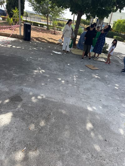 The image depicts an outdoor scene on a paved area, likely a public space given the presence of a trash can and warning tape. In the background, there are trees and structures that suggest an urban or suburban environment. The ground is concrete, with scattered shadows from overhead foliage, and several cigarette butts are visible.
In the mid-ground, a group of people is gathered. On the far left, a woman in white pants and a white jacket stands holding a white bag. To her right, a woman in a green dress is being helped by another person. A young boy in a black t-shirt and shorts is standing next to them. Further to the right, a woman in a dark blue patterned dress is drinking from a water bottle. A young girl in a white t-shirt and shorts stands on the far right of this group, looking towards the viewer. Some cardboard is laid out on the ground, possibly for seating or as a surface for an activity. The overall impression is a casual gathering in a somewhat unkempt outdoor setting.
While the image is not explicitly stated to be in Moraleda de Zafayona, Spain, the presence of specific architectural styles or vegetation, if identifiable, could provide clues. However, based solely on the visual information, a definitive location within Moraleda de Zafayona cannot be confirmed. The scene appears to be taking place during daylight hours, under a clear or partly cloudy sky, as indicated by the strong shadows.