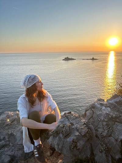 A woman sits on a rocky cliff overlooking the sea at sunset in Kaluđerac, Montenegro. The sun is visible on the horizon, with a bright reflection on the water. Two small rocky islands are visible in the distance. The woman is wearing a white shirt, green pants, and a white headscarf. She is looking out at the sea. She is wearing brown adidas shoes with white stripes.