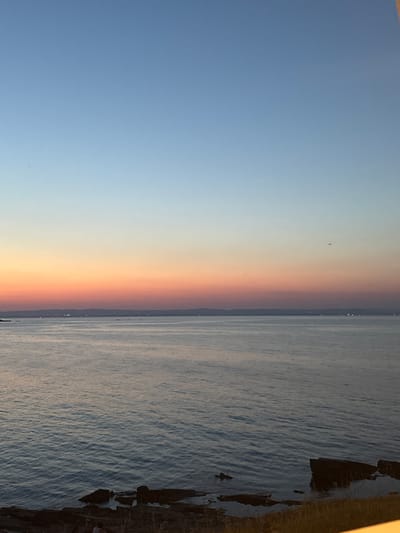 The image depicts a serene coastal scene at dusk. The sky transitions from a clear, pale blue at the top to a gradient of soft orange and pink hues near the horizon, indicating the setting sun. A body of calm water, likely the sea, occupies the lower half of the frame, reflecting the colors of the sky. In the distance, a landmass with faint lights suggests a town or city. The foreground features a rocky shoreline with some sparse vegetation. A single, distant object, possibly an aircraft, is visible in the sky. The scene is located in Chernomorets, Bulgaria, a coastal town known for its beaches and natural beauty.