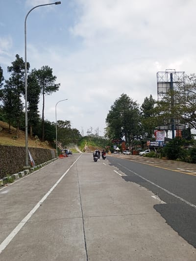 The image presents an uphill road in Ciloto, Indonesia, under a partly cloudy sky. The road's left side features a concrete surface, delineated by a continuous white line, a white and black curb, and a low stone retaining wall. This wall supports a grass embankment populated by tall evergreen trees. A prominent grey metal street light pole stands near the left edge, with a second pole visible further back. A small, multi-colored flag is attached to a pole on the left shoulder, where orange traffic barriers and a small parked vehicle or vendor stall are present in the midground. The right side of the road consists of an asphalt surface with white and yellow markings. Three individuals on motorcycles, wearing helmets, are proceeding uphill in the rightmost lane. On the right shoulder, two white cars are parked near a building complex. A sign for "POS POLISI PREDIKSI" is visible, along with another red and white sign above it. To the far right, a large metal billboard structure is partially obscured by trees. Dense vegetation and trees line both sides of the road and extend into the distant background.