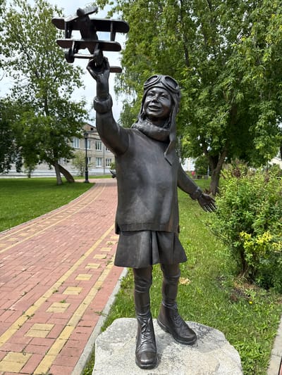 The image depicts a bronze statue of a young girl in aviator goggles and a scarf, holding a model airplane aloft. The statue is situated outdoors on a stone pedestal in a park-like setting. A red brick pathway with yellow accents is visible to the left of the statue, bordered by a manicured lawn. In the background, trees with lush green foliage are present, along with a light-colored building and a lamppost. The statue appears to be located in the city of Krasnoufimsk, Russia, specifically in a public space such as a park or square. The overall scene conveys a sense of youthful aspiration and dreams of flight.