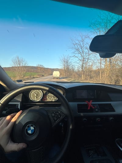 The image provides a first-person view from the driver's seat of a BMW E60 series vehicle, traveling on a paved rural road near Erden, Bulgaria. The driver's left hand is positioned on the black leather steering wheel, which features a prominent BMW emblem, carbon fiber trim, and integrated controls. The instrument cluster displays a vehicle speed of approximately 75-80 km/h, an engine speed of approximately 1500 RPM, an external temperature of 24°C, and an odometer reading of approximately 304,383 kilometers. A red, "X"-shaped object is clipped onto the central air conditioning vent on the dashboard.

Through the windshield, a white cargo van, consistent with a Mercedes-Benz Sprinter model, is visible ahead, proceeding in the same direction on the two-lane asphalt road. The roadside vegetation primarily consists of bare, leafless deciduous trees and bushes, indicative of late winter or early spring. In the background, rolling hills are visible, some covered with light green foliage, others with sparse, bare trees. The sky is clear blue, and the scene is illuminated by direct, low-angle sunlight, casting shadows. Small dark specks, possibly insects or debris, are discernible on the windshield and in the distant sky.