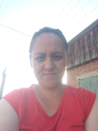 A woman with dark hair pulled back is looking directly at the camera in a close-up shot. She is wearing a red v-neck shirt. Behind her, a wooden fence with a wire mesh is visible, and further back, a brick building with a green door can be seen. The sky is bright and blown out, suggesting it is daytime. The image appears to be taken outdoors, possibly in a rural or suburban setting within the city of Tolbazy, Russia.
