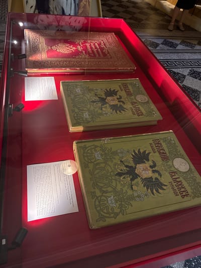 The image shows a display case with three antique books. The books are large and appear to be elaborately decorated. The central and rightmost books have green covers with a prominent two-headed eagle emblem and decorative floral motifs. The text on these covers appears to be in Cyrillic script. The leftmost book has a red cover with intricate gold embossed designs, also featuring an eagle emblem.
A white placard is positioned in front of the central book, containing text in Cyrillic. The placard provides information about the books, identifying them as "The Coronation Collection" celebrating the coronation of Emperor Nicholas II. It mentions that the collection was compiled under the direction of V.S. Krivenko and published by the Russian Empire, Saint Petersburg, in 1899. The text details the content of the volumes, which include historical accounts of Russian coronations, specific details of Nicholas II's coronation on May 4, 1896, and accompanying materials such as manifests, decrees, programs, and guest lists. It also highlights the rich illustrative content, including photographs, engravings, and works by various artists.
The display case is red, and the background behind the books shows a patterned floor with geometric designs. The overall impression is that of a museum exhibit showcasing historical and artistic artifacts.