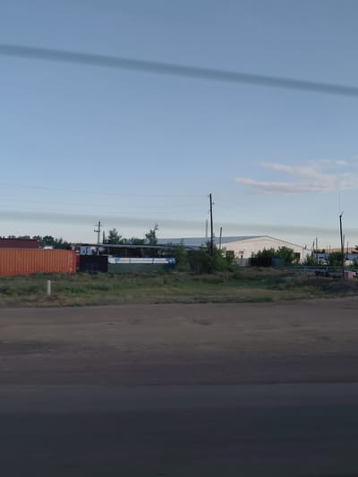 The image shows a roadside view in the vicinity of Молодёжный, Kazakhstan. In the foreground, a dirt road or path is visible, transitioning into a darker, smoother surface that appears to be a paved road or asphalt. Behind this, a strip of dry, brown grass and some sparse green vegetation lines the edge of the road.
Further back, there are several structures. To the left, two large, orange shipping containers are present, with one partially visible. Adjacent to the containers, there is a long, low building with a green and white corrugated metal exterior, partially covered by vegetation. To the right of this, a larger, white building with a sloped roof is prominent. This building appears to be a warehouse or industrial structure, with trees and bushes surrounding it.
Power lines and poles are visible across the scene, indicating infrastructure. The sky is a light blue with some wispy clouds and a prominent, dark horizontal shadow across the upper portion of the frame, likely cast by a power line or overhead structure. The overall impression is an industrial or semi-rural area on the outskirts of a settlement.