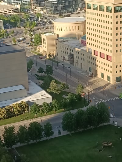 The image shows a view of the Square One area in Mississauga, Canada, from an elevated perspective. In the foreground, a well-manicured green lawn features a circular seating area with a fire pit and several small sculptures or decorative elements scattered across the grass. A line of mature trees with dense green foliage borders the lawn.
Beyond the lawn, a road runs parallel to the trees, with several cars parked along it and some moving on the street. Traffic lights and streetlights are visible along the road. A pedestrian walkway is situated between the trees and the road.
The midground of the image is dominated by a cluster of modern urban buildings. A tall, beige-colored building with numerous regularly spaced windows stands prominently, featuring a distinctive architectural style with protruding vertical elements. Adjacent to it, a circular building with arched window-like openings is visible. Another lower, beige building with a modern design is also present.
In the background, more urban structures can be seen, including what appears to be a multi-story parking garage and other contemporary buildings, suggesting a bustling downtown or commercial district. The overall scene depicts a city environment with a mix of green spaces and modern architecture, likely captured during daylight hours given the shadows cast by the buildings and trees.