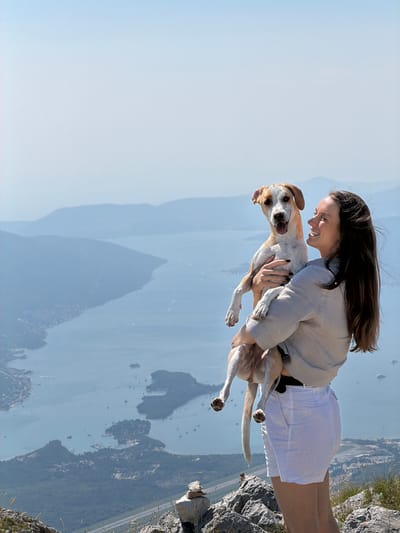 A young woman with long, dark hair and a light-colored shirt and white shorts holds a medium-sized dog with tan and white markings. The dog is looking forward with its mouth open and tongue out, appearing happy. They are standing on a rocky incline overlooking a vast body of water, possibly a bay or fjord, dotted with islands and boats. In the distance, hazy mountains rise up, layered one behind the other. The sky is a pale blue, suggesting a clear, sunny day. The scene appears to be in the vicinity of Kuk, Montenegro, given the distinctive mountainous coastal landscape and the presence of a settlement visible along the shoreline.