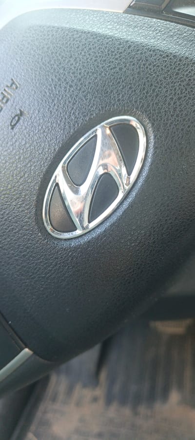 This is a close-up, angled shot of a Hyundai logo on a car's steering wheel. The logo is a silver, oval-shaped emblem with a stylized "H" inside, set against a textured black background that appears to be leather. The steering wheel itself is black and shows a pebbled texture. To the upper left of the logo, faint white text is visible, appearing to read "AIRBAG". The lighting suggests it is daytime, possibly indoors or in a shaded outdoor area, given the reflections on the metallic logo. The background below the steering wheel is out of focus and shows a blurred, dusty surface with vertical lines, possibly the floor mats or lower interior of the car. There are no people visible. The context provided indicates the location is Sosnoviy Bor, Russia.
