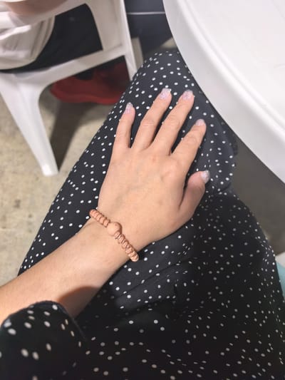 A close-up, high-angle shot captures a person's hand resting on their lap, which is covered by a black polka-dot skirt. The person's nails are painted with a sparkly, light pink polish. A thin, light brown bracelet, made of interconnected beads, is worn on their wrist. In the background, a white plastic chair is visible, with hints of red shoes underneath. The scene is set in an outdoor or semi-outdoor location, possibly a cafe or patio, with a concrete-like floor. The location is Белоярская Застава, Russia.