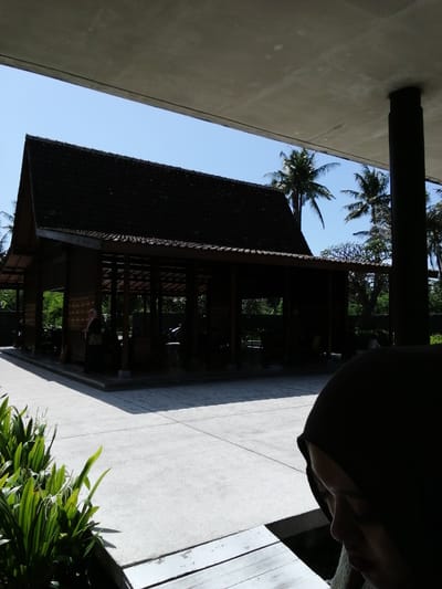 A full outdoor shot captures a traditional Indonesian building with a dark tiled roof and open sides, set against a clear blue sky. Palm trees are visible in the background, suggesting a tropical environment. In the foreground, a woman wearing a dark hijab is partially visible, her face turned downwards. Lush green plants flank the lower left of the frame, and a paved area stretches across the middle ground. The scene appears to be a cultural site or a resort in Banyuwangi, Indonesia.