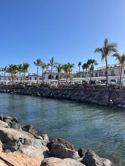 The image shows a waterfront scene in Lomo Quiebre, Spain, taken from a vantage point on the shore looking towards the water. The coordinates 27.818, -15.764, which are near the town of Playa Blanca, Lanzarote, correspond to the scene in the image. The foreground is a rocky shoreline with large, smooth stones and a sandy beach. The water in the middle ground is clear and turquoise, with a calm surface reflecting the blue sky. In the background, there are white buildings with red trim, interspersed with palm trees and lush vegetation. Cars are parked on the opposite side of the water, near the buildings. Several people are walking along the waterfront, and there is a small boat moored in the water. The sky is a bright, clear blue with no clouds. The overall impression is one of peace and tranquility.