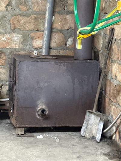 The image depicts a rustic scene in what appears to be an outdoor or semi-outdoor storage area, possibly in the city of Ayet, Kazakhstan. Dominating the foreground is a large, dark, rectangular metal stove, likely a wood-burning stove, showing signs of age and use. It rests on a concrete floor and is positioned against a brick wall. A metal pipe extends vertically from the top of the stove, suggesting a chimney system.
To the right of the stove, a metal shovel with a worn, grey scoop is leaning against the brick wall. The shovel handle is partially obscured by the stove. Above and behind the stove, a collection of green and yellow hoses are bundled together, secured with a yellow clip. Another metal pipe, wider and lighter in color than the chimney pipe, runs vertically up the brick wall to the left of the stove.
The brick wall itself is constructed from rough, weathered bricks in shades of brown and orange, interspersed with grey mortar. The concrete floor is uneven and shows some debris, including a crumpled piece of white paper. The lighting suggests an overcast day or an indoor space with limited natural light. There are no people or animals visible in the image. The overall impression is one of a utilitarian and somewhat disheveled workspace or storage area.