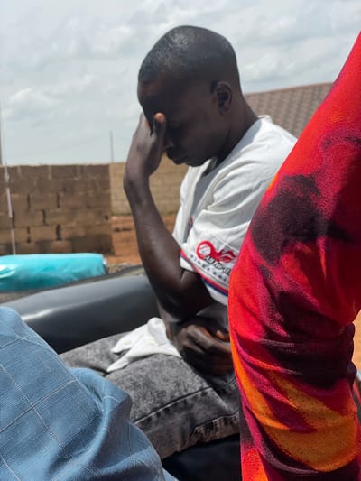 In the city of Kassa, Nigeria, a man with dark skin and short hair sits with his head bowed, his right hand pressed against his forehead. He is wearing a white t-shirt with a circular logo on the left side and a white cloth draped over his lap. To his right, a portion of a person's arm is visible, adorned with a red and orange tie-dye patterned fabric. The background consists of a brick wall and a partially visible white and blue object. The man appears to be in a contemplative or distressed state, possibly outdoors given the bright sky and the general outdoor ambiance suggested by the brick wall and open sky. The overall impression is a candid moment captured, focusing on the man's posture and the textures of the clothing.