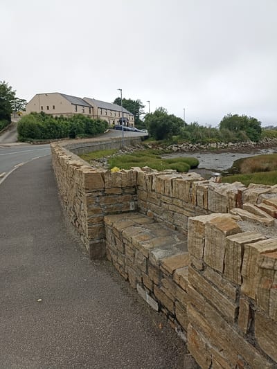 This image captures a scenic view of Annagry West, Ireland, under an overcast sky. The foreground is dominated by a rustic stone wall, meticulously constructed with rough-hewn, light-brown stones. The wall follows the curve of an asphalt road, which leads away from the viewer and into the distance. To the left of the road, a grassy embankment is visible.

Beyond the road and the wall, the scene opens up to a tidal estuary with mudflats, bordered by lush green vegetation and reeds. In the mid-ground, a long, beige building with a dark roof stands prominently, identified by visible text as a "HOTEL." Several parked cars are visible in front of the hotel, suggesting it is open for business. A streetlamp stands near the hotel entrance.

The overall atmosphere is one of tranquility and natural beauty, enhanced by the muted light of the cloudy day. The presence of the hotel hints at tourism in the area. The natural landscape of the estuary and the traditional stone wall evoke a sense of place specific to coastal Ireland. There are no people visible in the image. The time of day appears to be daytime, given the ambient light.