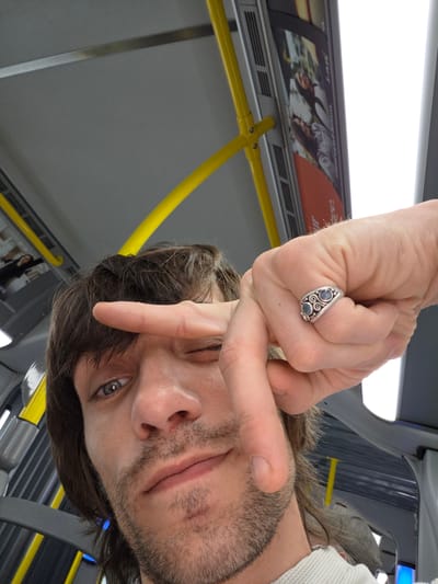 The media file shows a close-up selfie taken from a low angle inside a vehicle, likely a bus, as indicated by the yellow grab handles and the interior ceiling. The subject is a person with dark, medium-length hair and stubble. They are looking towards the camera with one eye partially closed, and their right hand is raised, with two fingers extended in a peace sign gesture. The person is wearing a silver ring with two blue stones on their right ring finger. The lighting suggests it is daytime. In the background, advertisements are visible on the interior wall of the vehicle, one featuring two women. The overall mood appears casual and informal.