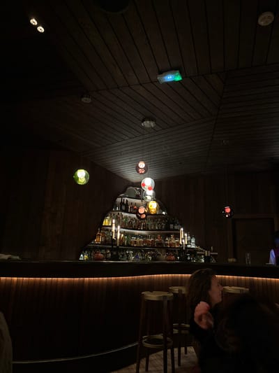 This media file captures the interior of a dimly lit, stylish establishment, likely a bar or lounge, with a warm and intimate atmosphere. The image is rotated 90 degrees clockwise.
The scene features a modern design dominated by dark wooden paneling on the ceiling and walls, arranged in a striking geometric pattern of angled planks. Scattered across this wooden surface are various light fixtures: several small, recessed spotlights, a circular smoke detector-like device, and a rectangular emergency light or control panel emitting a green glow.
A central element of the visual composition is a cluster of modern pendant lights hanging at varying heights. These lights feature spherical glass shades in a diverse palette of colors including red, amber, clear, green, and a muted brown or grey, casting soft, colorful glows.
On the right side of the image (which would be the floor and wall if the image were correctly oriented), a well-stocked bar is visible. Numerous bottles of spirits and liqueurs are neatly arranged on shelves, illuminated from within or by overhead lights, creating bright reflections. The bar counter itself features a contemporary design with a warm, illuminated strip running along its length. Several elegant bar stools with dark seats and light-colored bases are positioned in front of the counter.
One person is visible on the far right, seated at or standing near the bar. Only their head and upper torso are visible in profile, showing dark, possibly curly hair. They appear to be looking forward or slightly downward, suggesting a moment of quiet contemplation or waiting.
The overall impression is one of a sophisticated and cozy evening setting, characteristic of a high-end lounge or restaurant. The dim, colorful lighting and dark wood create an inviting ambiance. No specific activity beyond being present at the bar is distinctly captured, and no discernible text is visible within the frame. The time of day is clearly evening or night due to the reliance on artificial lighting. The location context indicates Val-d'Isère, France.