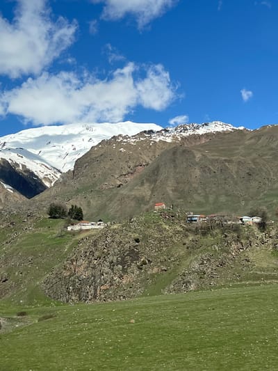 The image shows a mountainous landscape in Sioni, Georgia, with a snow-capped peak on the left side of the frame. Patches of snow are scattered along the mountain's ridge. To the right, a steep, rocky hillside with sparse vegetation descends to a green meadow. A small village is situated on the slope. The sky is blue with scattered white clouds.