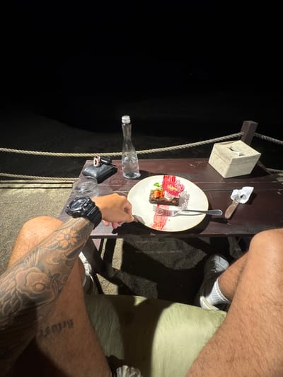 The image presents an overhead view of a partially illuminated outdoor setting. A dark background suggests nighttime or a dimly lit area. A person wearing a black wristwatch on their left arm, which features prominent tattoos, is holding a fork and appears to be eating a dessert on a white plate with a red decorative element. The dessert appears to be a slice of cake with a strawberry. A knife rests near the plate. A tissue box sits near the plate, and there are two empty glass bottles on the side of the table. Another person wearing white sneakers, socks, and light-colored shorts sits with their legs and thighs within the frame. The setting appears to be a wooden table that is a part of a structure supported by ropes and beams, which could be part of a restaurant, bar, or some other outdoor venue, that is consistent with the city of Munggu, Indonesia.