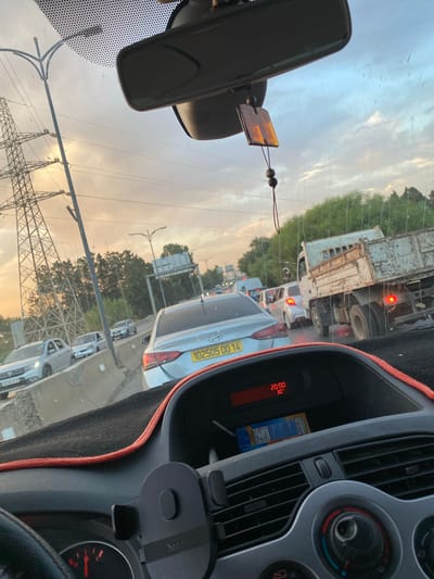 The image captures a congested roadway in El Karia, Algeria, during what appears to be late afternoon or early evening. The perspective is from inside a vehicle, looking forward through the windshield. Several cars are visible on the road, all seemingly stopped or moving very slowly in traffic. A prominent power line pylon stands tall on the left side of the frame, with power lines stretching across the sky. The sky is partly cloudy, with hues of orange and yellow suggesting sunset.
In the foreground, the interior of the car is visible, including a dashboard with a digital display showing the time as "20:00" and a temperature of "30°C". A car phone holder is mounted on the dashboard. Behind the dashboard, a blue and yellow box is partially visible in a compartment.
On the road, a silver sedan with a license plate reading "102505 00 14" is in the center lane. To the right of this sedan, a light blue truck with a rusted bed is also in traffic. Several other vehicles are visible in the background, further indicating heavy traffic. Trees and vegetation line the side of the road, and a concrete barrier separates lanes of traffic. The overall scene depicts a typical traffic jam in an urban or suburban environment.